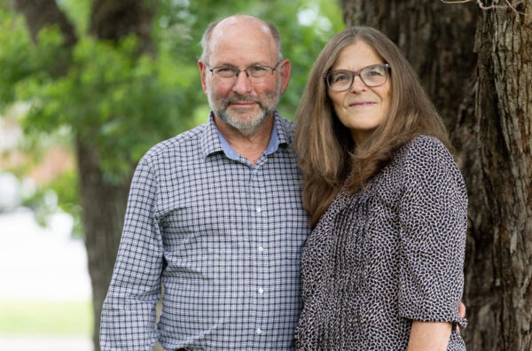 A man with black glasses and in a blue and white checkered shirt stands with a woman with long brown hair and glasses.