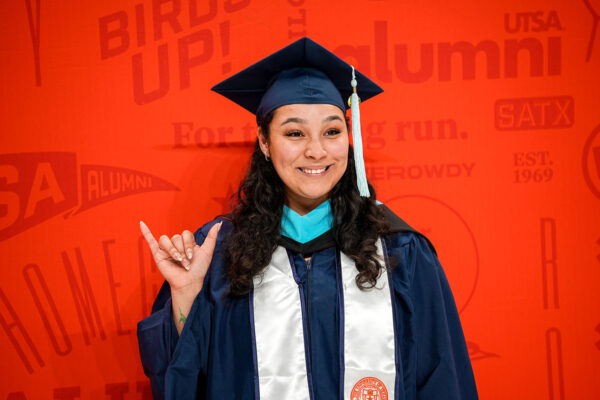 A woman in a navy blue graduate cap and gown stands against an orange background. She holds up her hand while making a hand gesture, which resembles a roadrunner.