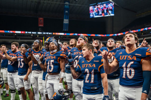 Dozens of football players in full gear stand on a field as they sing together.