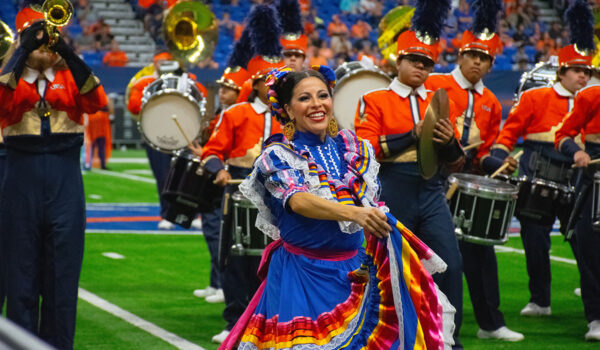 A woman dances in a colorful dress while the Spirit of San Antonio marching band performs on a football field