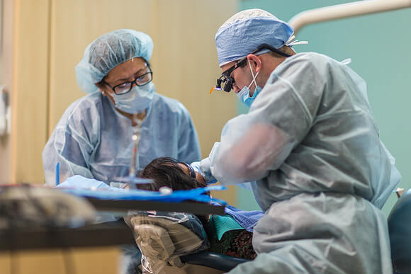 Two people are dresed in medical caps and gowns as they use instruments to check on someone's teeth.