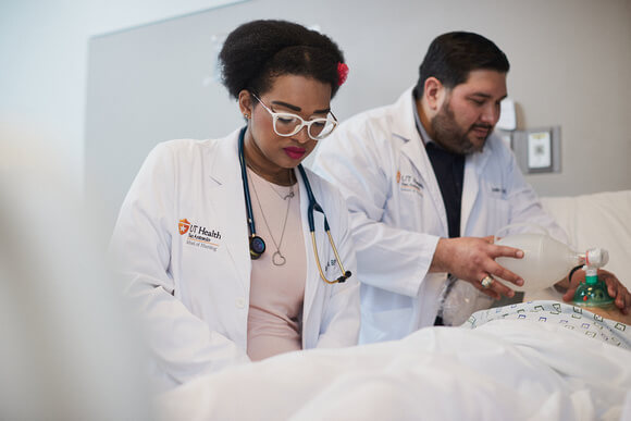 A woman and man are dressed in white doctor coats as they stand beside a patient laying down.