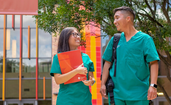 A woman and man in medical scrubs look at each other as they walk and speak.