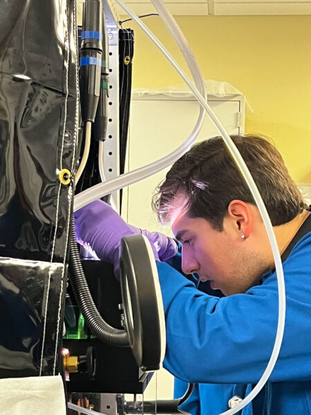 A man in a blue lab coat and purple gloves works on a piece of equipment with wires and tubes sticking out.