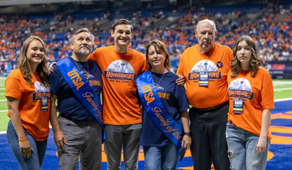 A family of six dressed in orange shirts smile as they take a photo together on a football field.