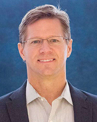 A man in glasses smiles at the camera in a grey jacket.