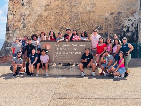 A group of young students gather around a sign that says, "San Juan National Historic Site."
