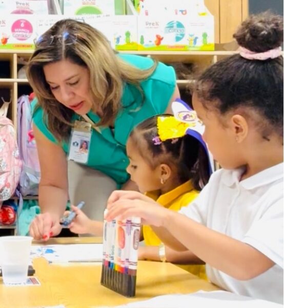 A woman in a blue shirt leans over a table with two girls and she helps them with their classwork.