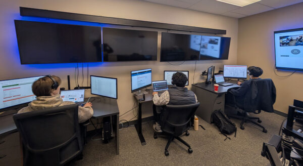 Three people are working in a control room with multiple screens and computers.