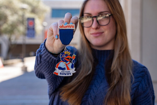 A blonde woman with glasses holds up the UT San Antonio Alumni Association fiesta medal.