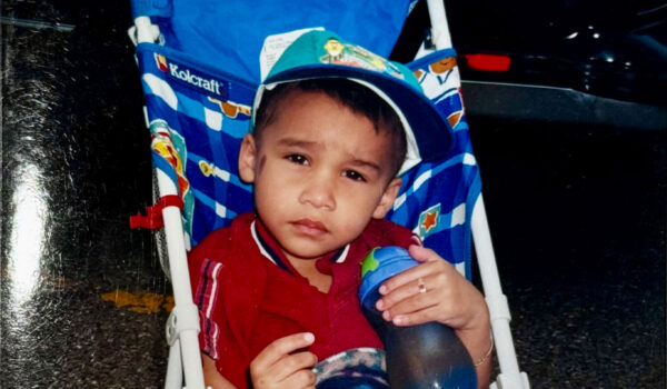 A young child sits in a colorful stroller, wearing a cap and holding a water bottle. 