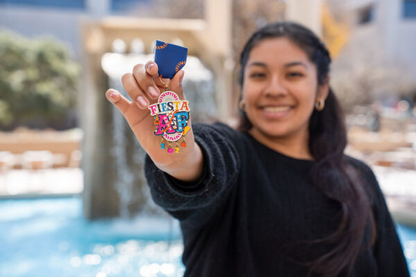 Smiling woman holds a colorful "Fiesta" medal with decorative elements, in front of a fountain.