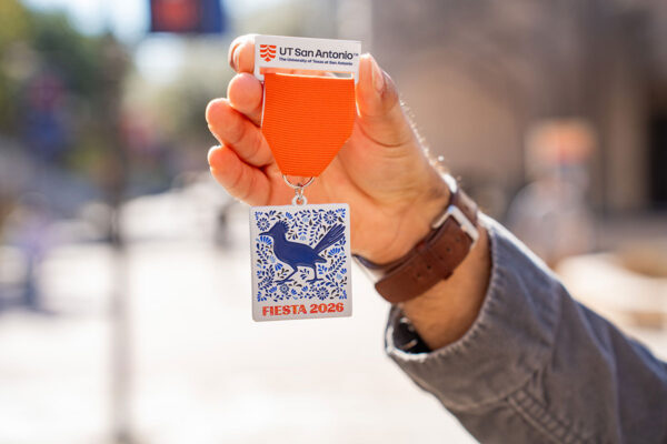 A hand holds an orange ribbon medal labeled "UT San Antonio." The medal features blue floral patterns, a bird, and the words "Fiesta 2026."