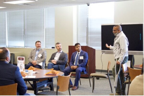 A man stands and gestures while speaking to three seated men in business attire during a meeting. 