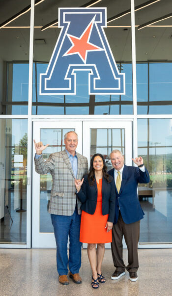 Taylor Eighmy, Lisa Campos and Mike Aresco put their "Birds Up" in front of a set of glass doors with the American Conference logo above it