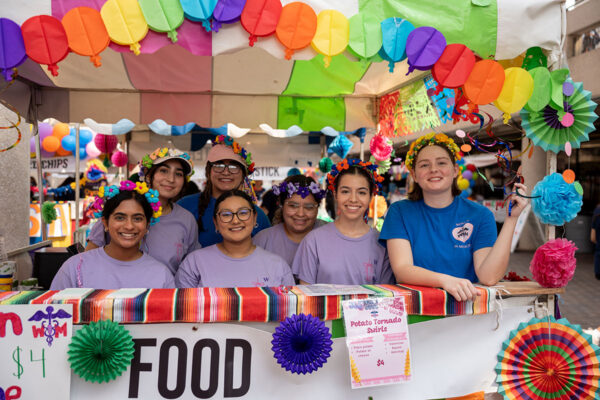 Seven students stand behind a colorful decorated food booth while they pose for a photo.