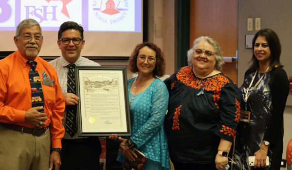 A group of five people stands together smiling, holding a framed certificate. They are in a room with a projector screen in the background