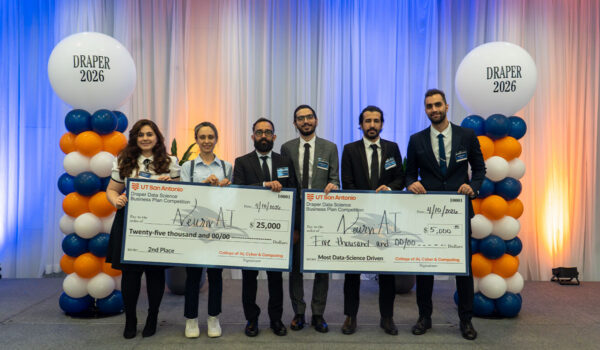 A group of six people stand smiling, holding large checks for a data science competition. Background features balloon columns labeled "Draper 2026."