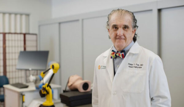 Elderly man in a lab coat and bow tie stands in a medical office. Background shows a robotic arm and medical equipment