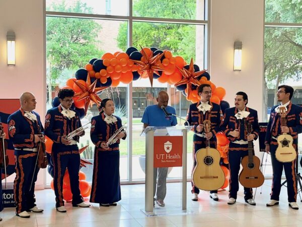A man at a blue shirt stands at a podium with a balloon arch behind him. Mariachi musicians stand on either side of him.