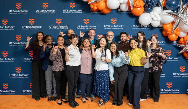 A group of smiling people pose with hand signs in front of a UT San Antonio backdrop and colorful balloons.