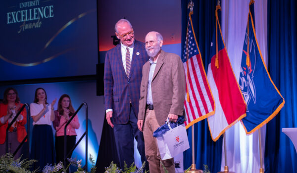 Two men stand on a stage during an award ceremony. One holds a gift bag with a logo.