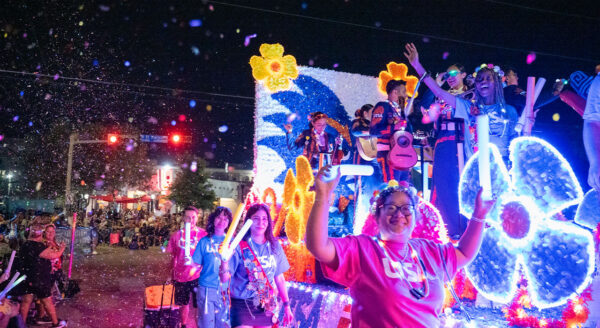 Three women hold up their hands waving as a lit up float passes beside them. Confetti is flying in the air as they go by.