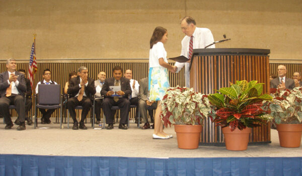 A young woman receives a certificate from a man at a podium during a ceremony. Seated people applaud, and potted plants with colorful leaves line the stage.