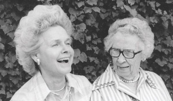 A black and white photo with two elderly women smiling warmly in front of a leafy background.