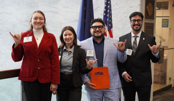 Four people stand side by side, smiling, holding up hand signs. One holds a folder labeled "UTSA." U.S. flags are visible in the background.