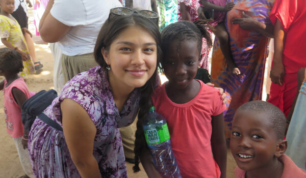 A smiling woman in a purple dress poses with two smiling children, one holding a water bottle.