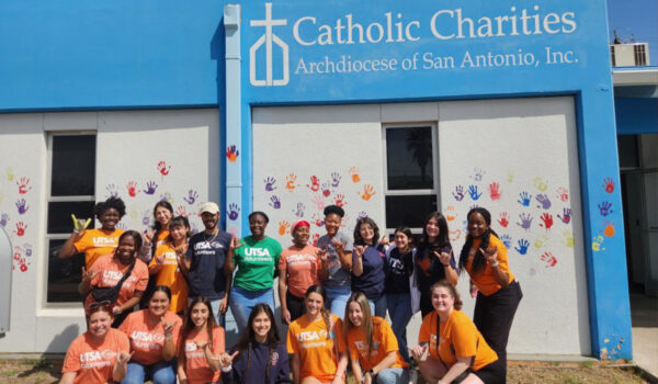 A group of smiling young volunteers in colorful UTSA shirts pose in front of a Catholic Charities building with handprints on the wall.