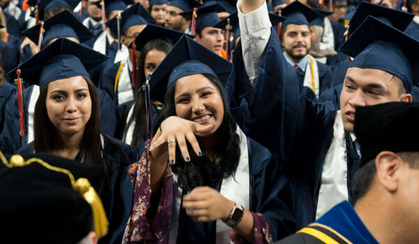 Graduates in blue caps and gowns celebrate at a ceremony. A smiling woman in the center shows her ring.