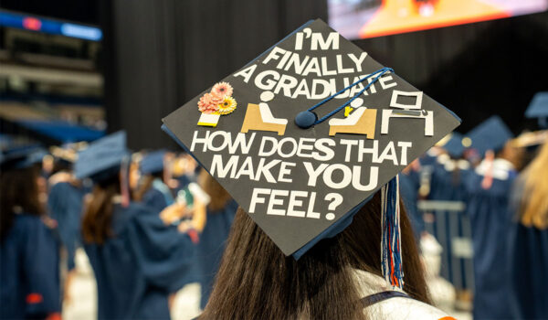 Graduation cap with creative design reading "I'm finally a graduate, how does that make you feel?" adorned with flower and computer icons. 