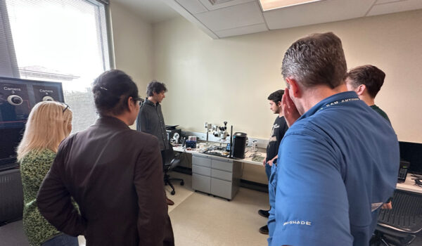 A group of six people stands in a room observe a desk filled with electronic devices.
