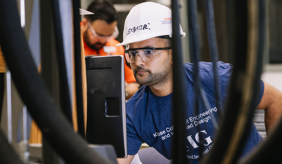 A male engineering student wears a hard hat while looking intently at a computer monitor.