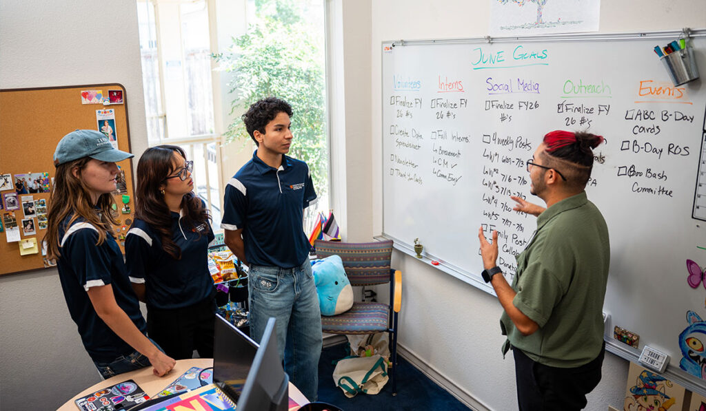 Three individuals in blue polos listen to another person with red and black hair as they point at a whiteboard.