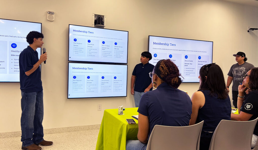 Two people stand next to TVs as they give a presentation in front of three people at a table.