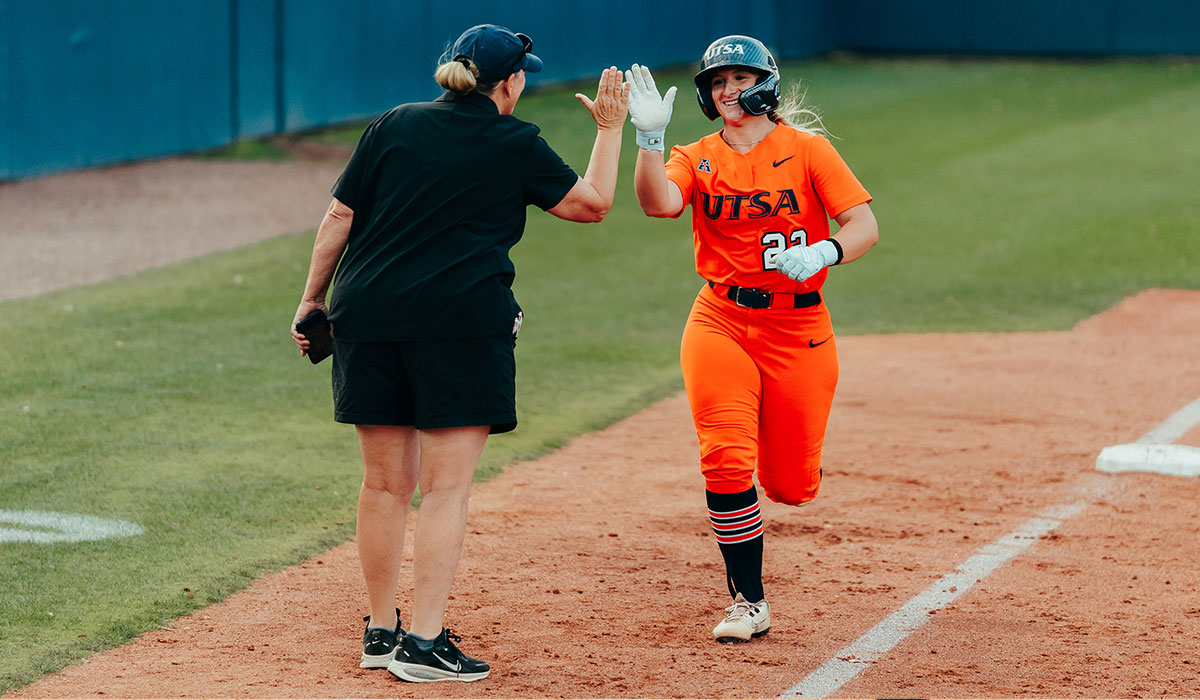 A woman in an orange softball uniform runs down the field to high-five a woman in a black athletic dress.