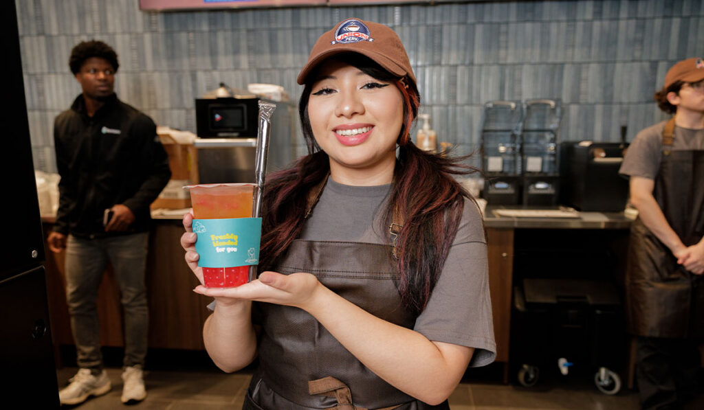 A barista in a ball cap and apron holds up a drink smiling with a coffee shop in the background.