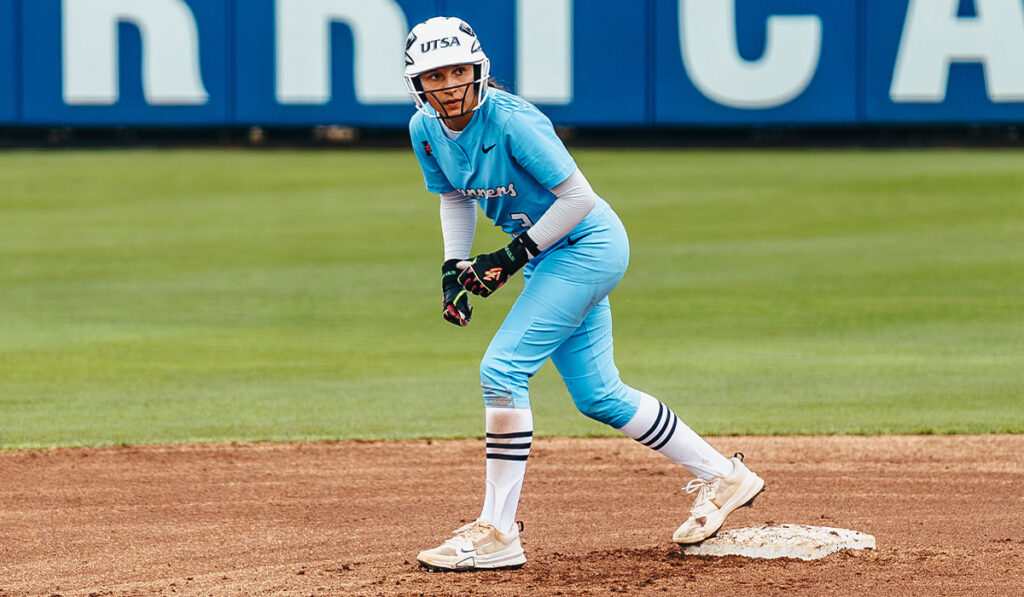 A woman in a baby blue softball uniform learns forward off the base.