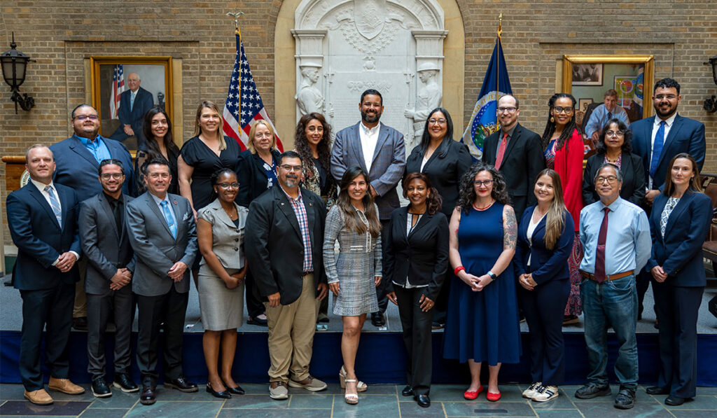 A group of professionals posing in a historic brick hall with American and state flags and framed portraits behind them.