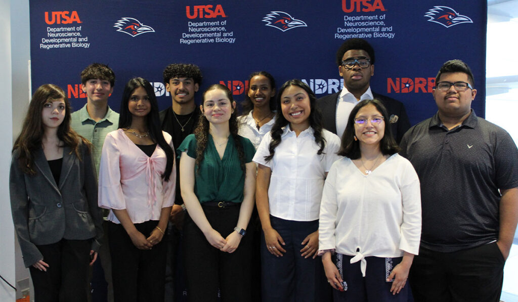 A group of students and faculty pose smiling before a UTSA neuroscience event backdrop featuring the Rowdy logo.