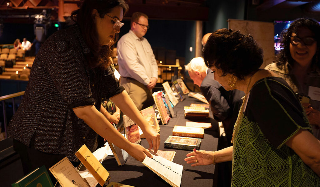 Two women browse books and chat across a display table while attendees gather in the background.