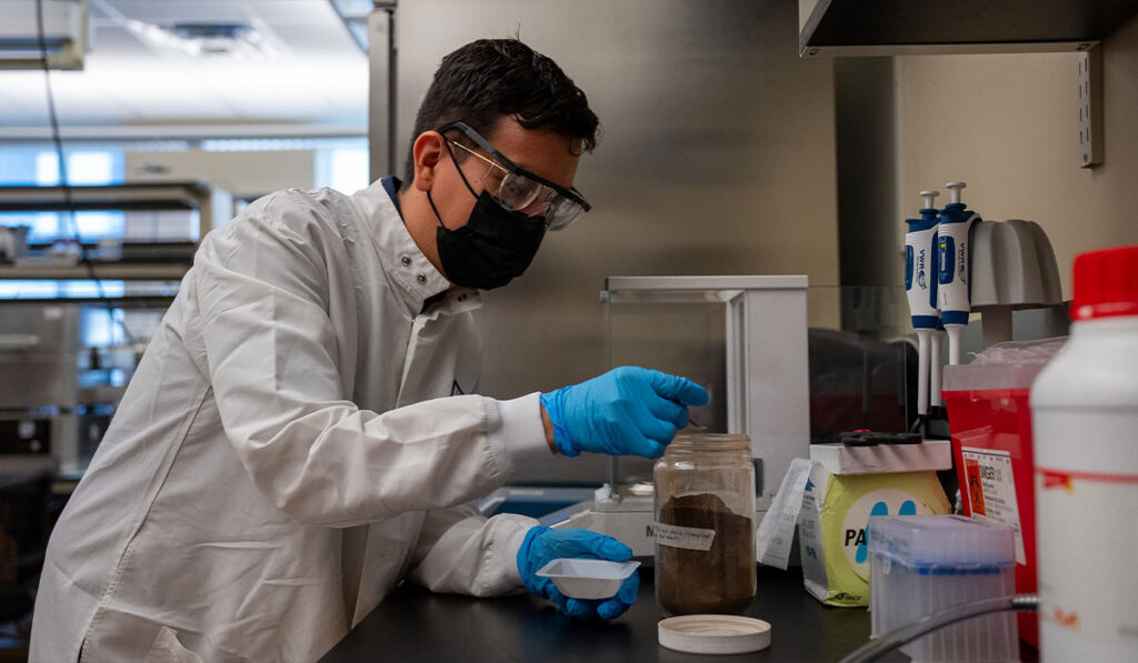 Masked laboratory scientist in a white coat and blue gloves handles samples at a bench.