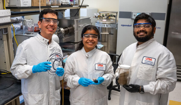 Three smiling researchers in protective lab suits, goggles, and gloves pose with equipment in a laboratory.