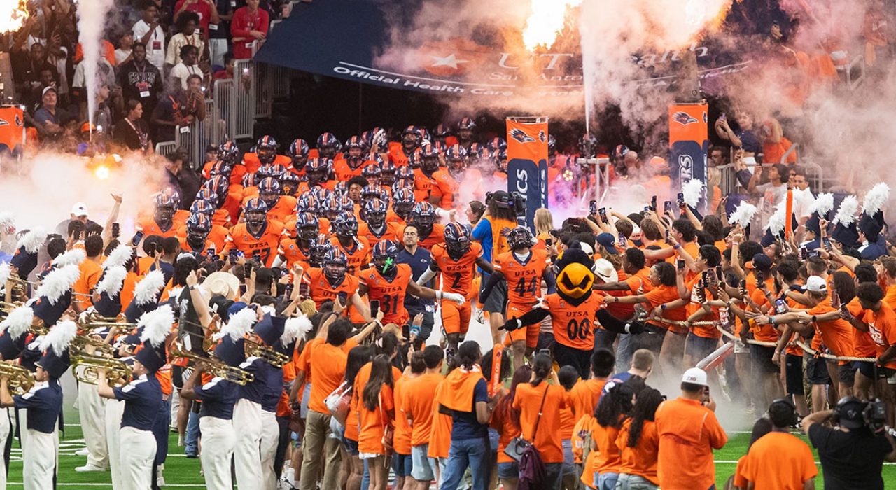The UTSA football team runs on to the Alamodome field while students in orange shirts cheer