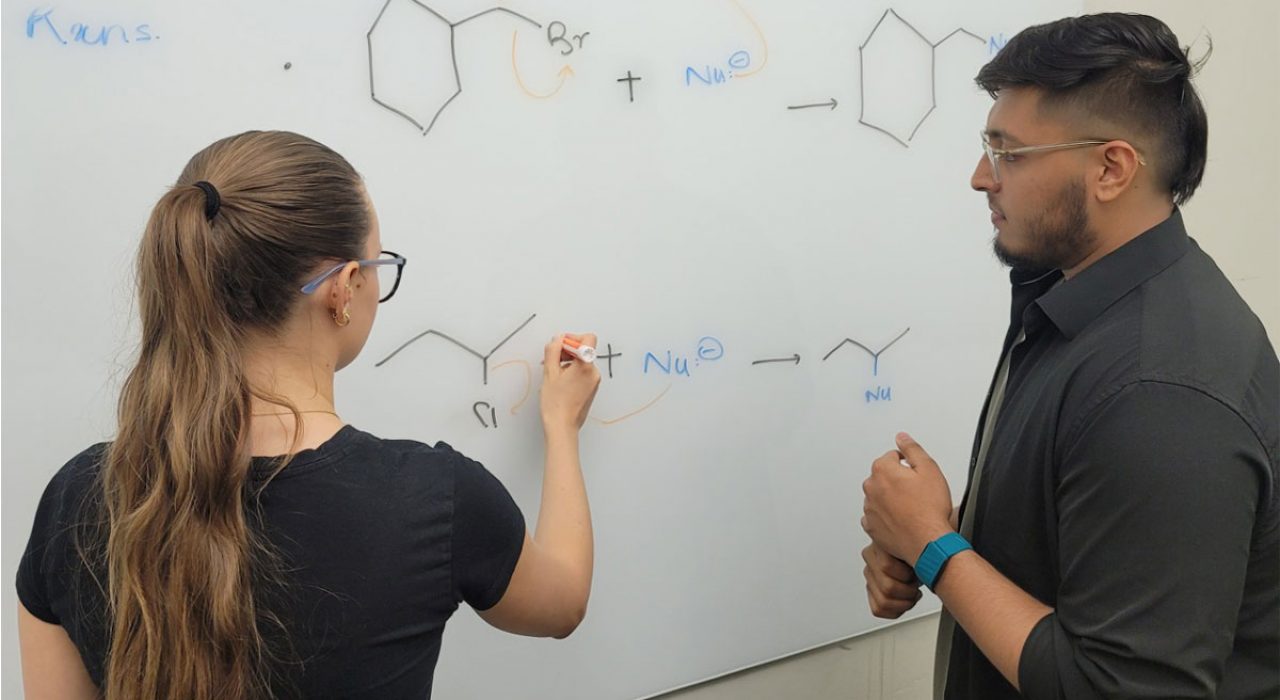 Two individuals in black shirts write out scientific formulas on a whiteboard.