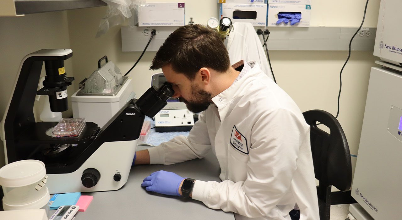 A man in a white lab coat looks into a microscope while conducting research in a lab.