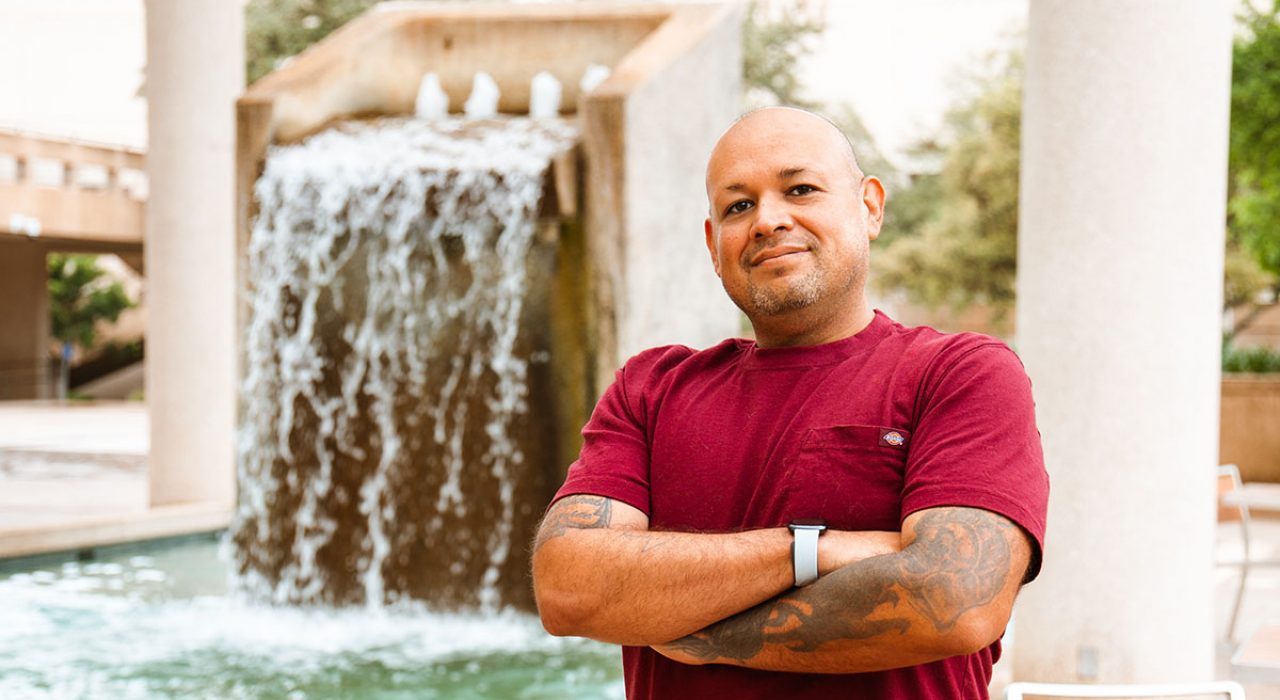 AJ Delgado stands with his arms folded next to the Sombrilla Fountain
